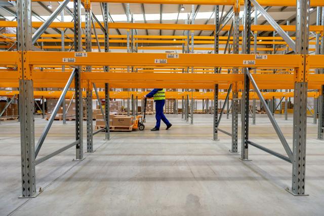 A worker moving a pallet jack down the aisle of a high-bay warehouse lined with empty pallet racking